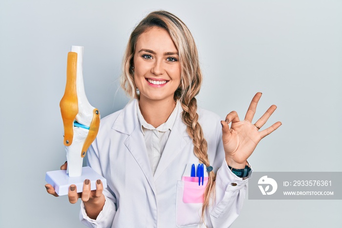 Beautiful young blonde doctor woman holding anatomical model of knee joint doing ok sign with fingers, smiling friendly gesturing excellent symbol