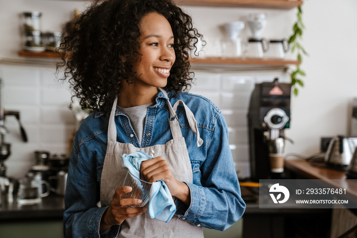 Black barista woman wearing apron wiping glasses while working in cafe