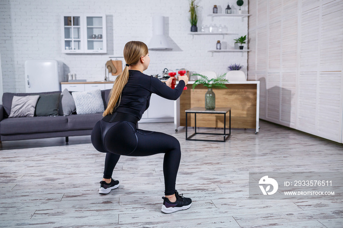 A slender young woman goes in for sports at home in a top and leggings. Fitness at home for a beautiful body. Exercises with dumbbells for the female body. Healthy lifestyle.