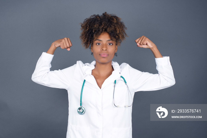 Waist up shot of caucasian doctor woman raises arms to show her muscles feels confident in victory, looks strong and independent, smiles positively at camera, stands against gray background.