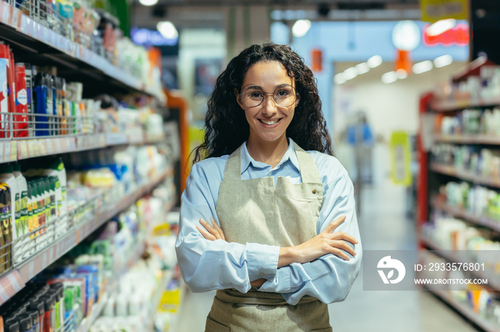 Portrait of Hispanic woman seller in supermarket, female worker in apron with curly hair and glasses smiling and looking at camera, saleswoman among goods with household chemicals and shelves.