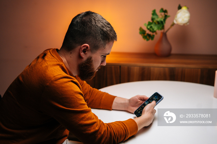 Close-up portrait of handsome young man using typing mobile phone sitting at table in dark living room at home. Closeup of bearded guy using smartphone alone.