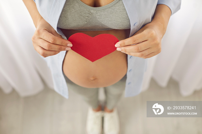 Close up crop top view of pregnant woman in pajama hold paper heart sign on belly show love and care to baby child. Healthy female excited with pregnancy. Motherhood and parenting concept.