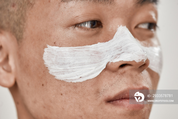 Close up of face of young asian man with problematic skin and hyperpigmentation applied mask on his face, looking away isolated over white background. Beauty, skincare routine