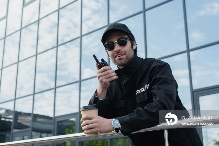 smiling guard with takeaway drink talking on walkie-talkie outdoors.