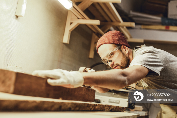 Attractive guy in protection goggles working with lumber plank on workbench in professional joinery