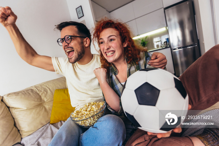 Happy young couple spending weekend together at home sitting on sofa watching soccer play live on tv and cheering for favourite team