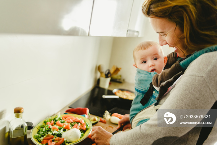 Busy mother preparing food in the kitchen while taking care of her baby, in a baby carrier using the kangaroo method.