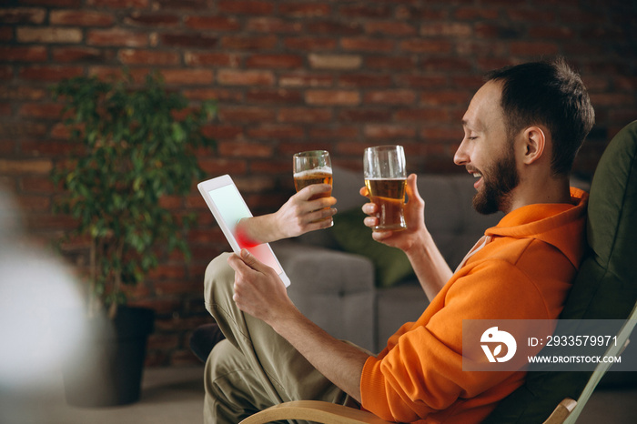 Happiness. Young man drinking beer during meeting friends on virtual video call. Distance online meeting, chat together on tablet at home. Concept of remote safe meetings and entertainment.