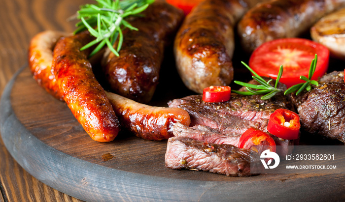 Close-up photo of mixed grilled meat platter. Beef, pork, poultry, sausages, grilled garlic, chili pepper, red tomatoes on wooden rustic background.