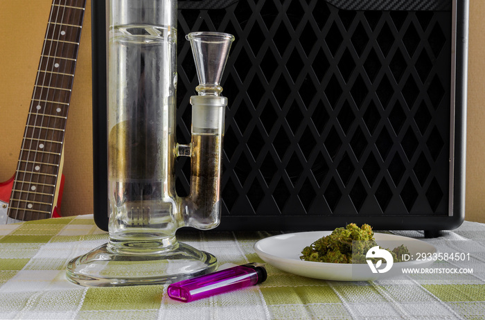 A transparent crystal bong weed, a plate full of marihuana and a purple lighter with a red electric guitar and amplifier in the background. Inspiring indoor photo.