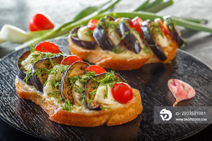 Bruschetta with fried eggplants, fresh tomatoes and cheese on black boards against the background of fresh vegetables. Close-up