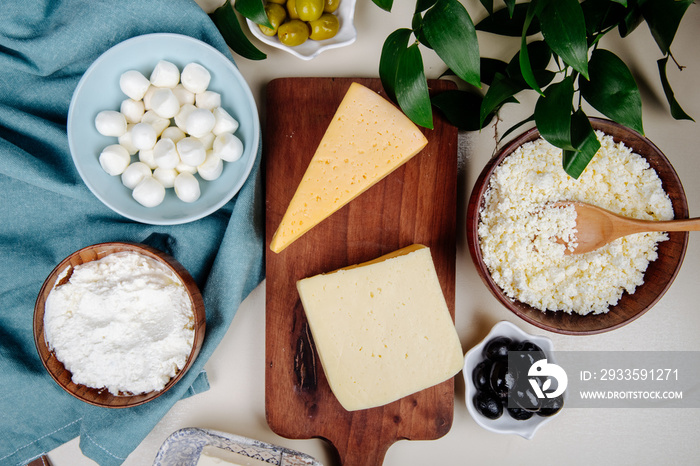 top view of different kind of cheese on wooden cutting board and cottage cheese in a wooden bowl with pickled olives on rustic background