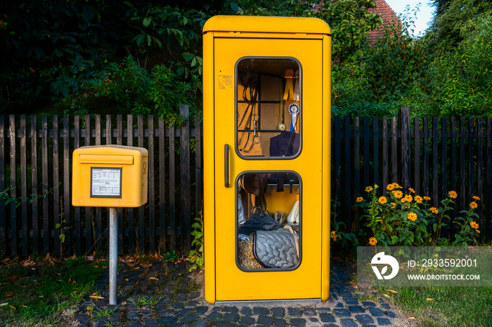 Ein gelber Briefkasten steht neben einer gelben Telefonzelle,  in der, weil sie nicht mehr funktionstauglich ist, Reitutensilien ausgestellt sind, im Hintergrund ein Holzzaun vor Bäumen und Büschen