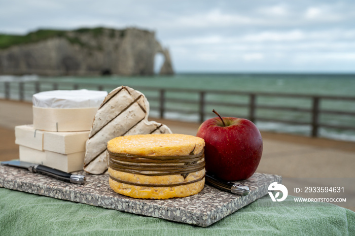 Cheeses of Normandy, round camembert cow cheese, yellow livarot, heartshaped neufchatel and view on alebaster cliffs Porte d’Aval in Etretat, Normandy, France