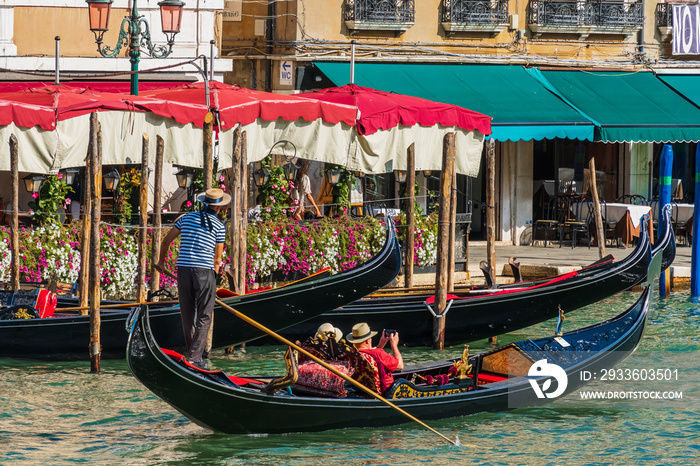 A gondolier standing in a gondola steers a boat with an oar in his hands, an elderly couple of tourists in a gondola filming a journey on smartphones along the coast of a Venice city street