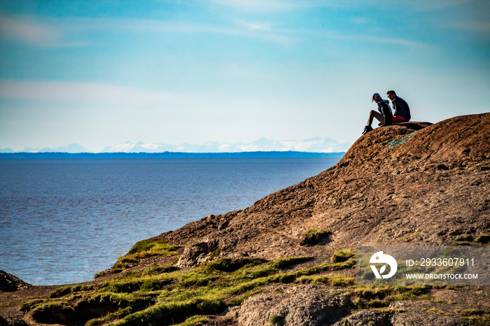 2 people sitting on the edge of a cliff over looking Cook inlet under a blue clear sky  near Anchorage, Alaska.