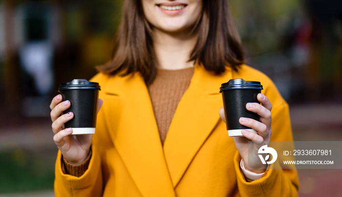 Smiling girl in a coat holding a black paper cup of coffee. Girl holding a paper cup with coffee or tea.