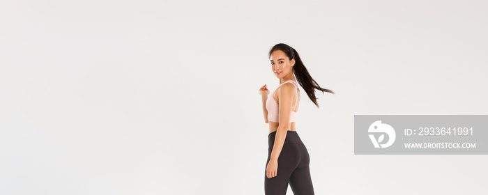 Full length of sassy good-looking, slim asian girl doing fitness, female athelte or workout coach walking with confident, motivated expression, turn at camera pleased smile, white background