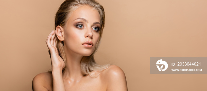 pretty woman with makeup on perfect face touching hair while looking away isolated on beige, banner