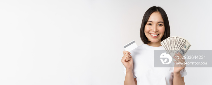 Portrait of asian woman smiling, holding credit card and money cash, dollars, standing in tshirt over white background