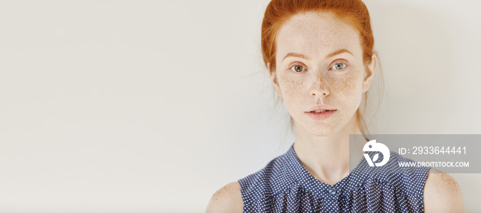 Eyes of different colors: blue and brown. Tender freckled young Caucasian female with heterochromia iridum wearing sleeveless shirt with spots having rest indoors, looking at camera with faint smile