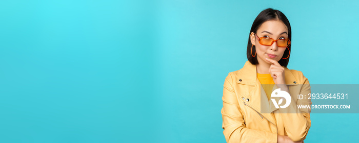 Portrait of asian woman thinking, looking thoughtful, searching ideas or solution, wearing sunglasses, standing over blue background