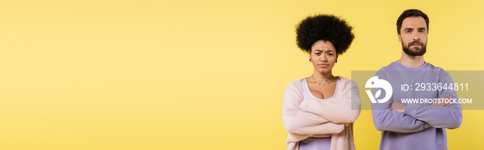 frustrated interracial couple looking at camera while standing with crossed arms isolated on yellow, banner.