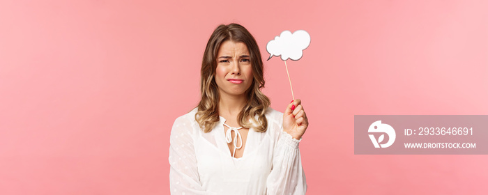 Spring, happiness and celebration concept. Close-up portrait of skeptical and hesitant young blond girl express dislike or reluctance, holding commend cloud stick near head, grimacing disappointed