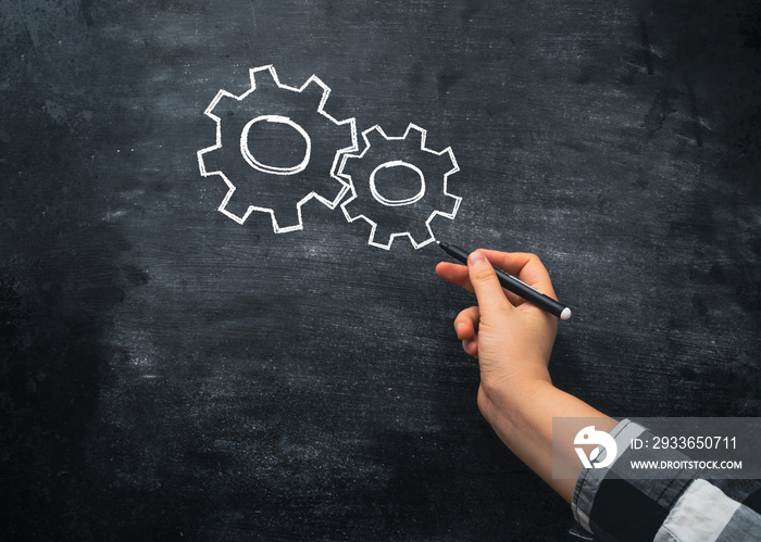 Person hand draws gears with white marker on blackboard