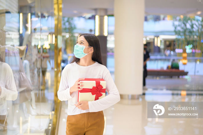 Woman wearing protective mask holding a gift box in shopping mall, shopping under Covid-19 pandemic,