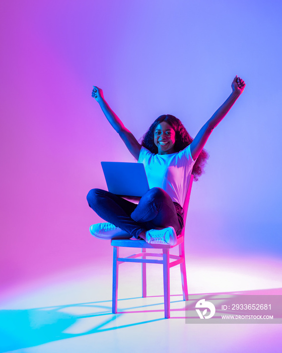 Excited young black lady sitting on chair with laptop, raising arms up, celebrating success in neon 