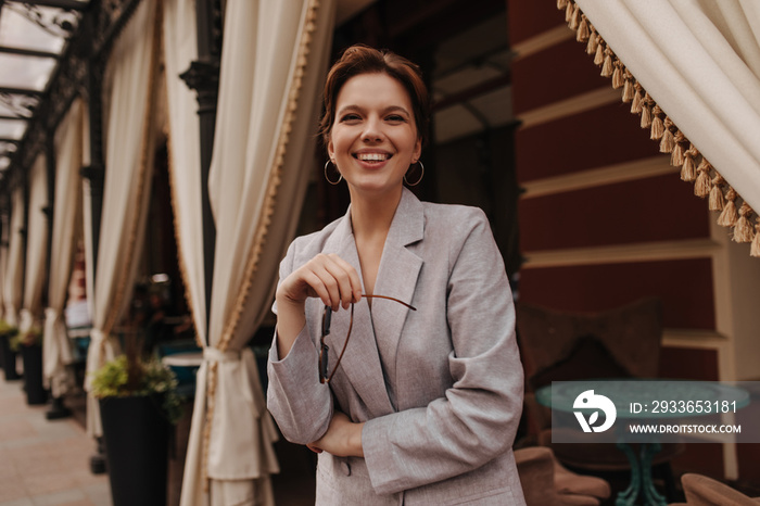 Woman in great mood looking into camera and posing near restaurant. Cheerful girl in grey oversize j