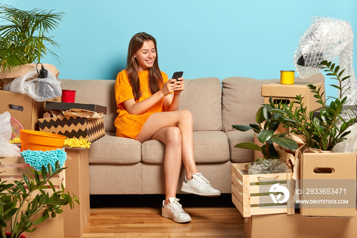 Moving concept. Cheerful young female model surrounded with lot of boxes, sits on comfortable sofa i