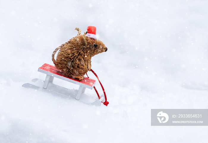 Cute little mouse hedgehog toy in red Christmas santa hat sledding on wooden sled from snow hill. Wi