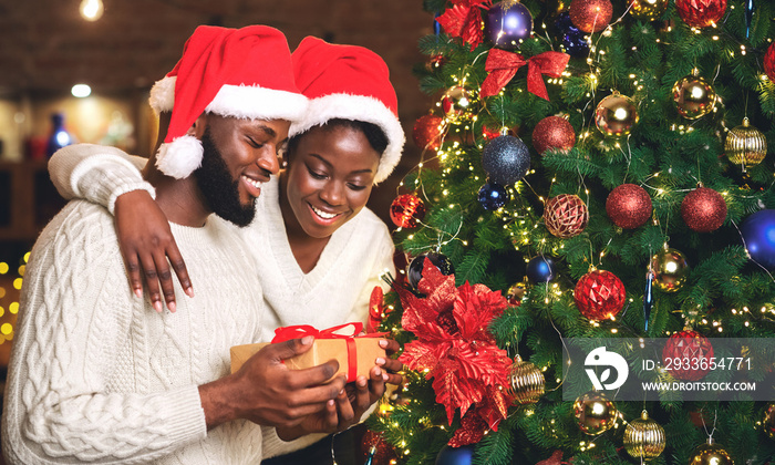 Portrait of happy afro couple holding gift and standing near Christmas tree