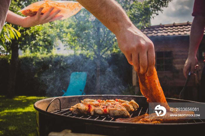 Hand of a person laying bacon steak  meat onto a smoker grill with other meat. Back yard garden BBQ 