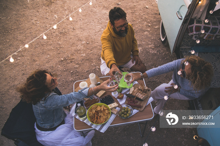 Happy friends cheering and drinking wine during camping vacation at the beach with vintage van. Peop