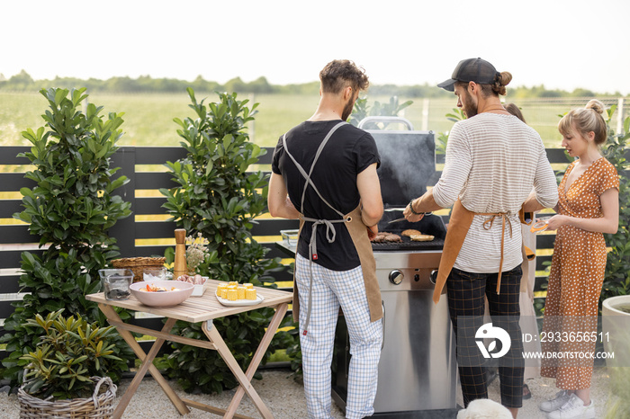 Happy young friends hanging out together, grilling food on a modern grill at beautiful backyard of a