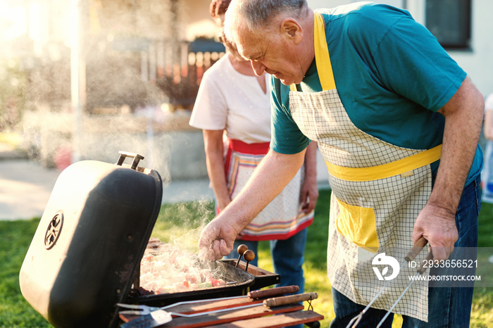 Grandparents with aprons grilling meat and vegetables on sticks while standing in backyard at spring