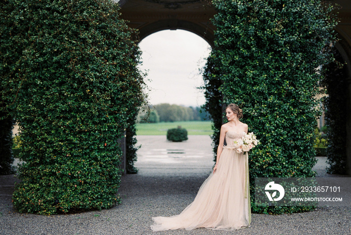 Bride with a bouquet stands near arches overgrown with green ivy. Como, Italy