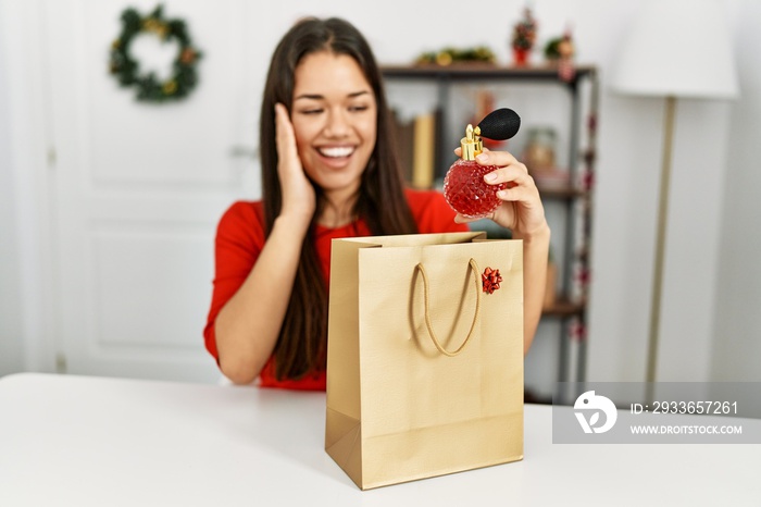 Young latin woman holding perfume of gift bag sitting by christmas decor at home