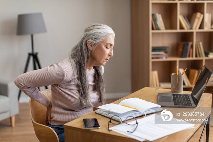 Unhappy caucasian old female with gray hair works on computer and suffers from back pain in room int