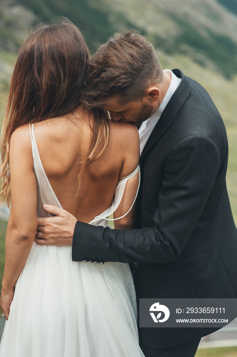 Groom in a dark suit kisses a beautiful brunette bride on her necked shoulder. Lifestyle, female bea