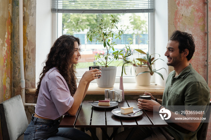 Young woman and her smiling boyfriend with cups of coffee sitting by table in front of one another a