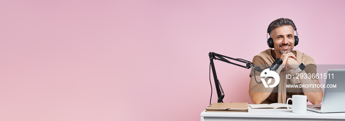 Handsome mature man in headphones using microphone while recording podcast against pink background