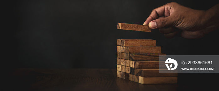 Close-up hand is placing wood block tower stacked in stair step with caution to prevent collapse or crash concepts of financial risk management and strategic planning.