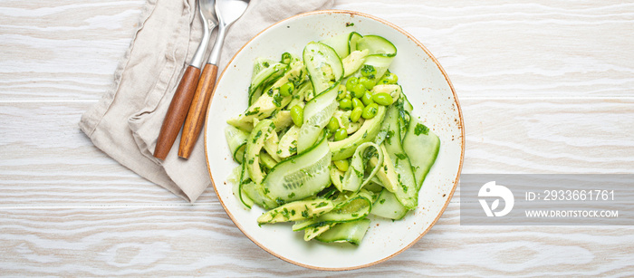 Healthy vegan green avocado salad bowl with sliced cucumbers, edamame beans, olive oil and herbs on ceramic plate top view on white wooden rustic table background