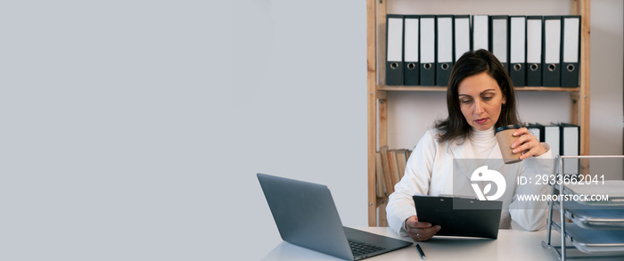 Tired woman doctor working in clinic office at table using laptop and clipboard drinking coffee