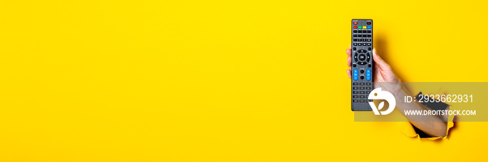 Female hand holding a TV remote control on a bright yellow background. Banner.